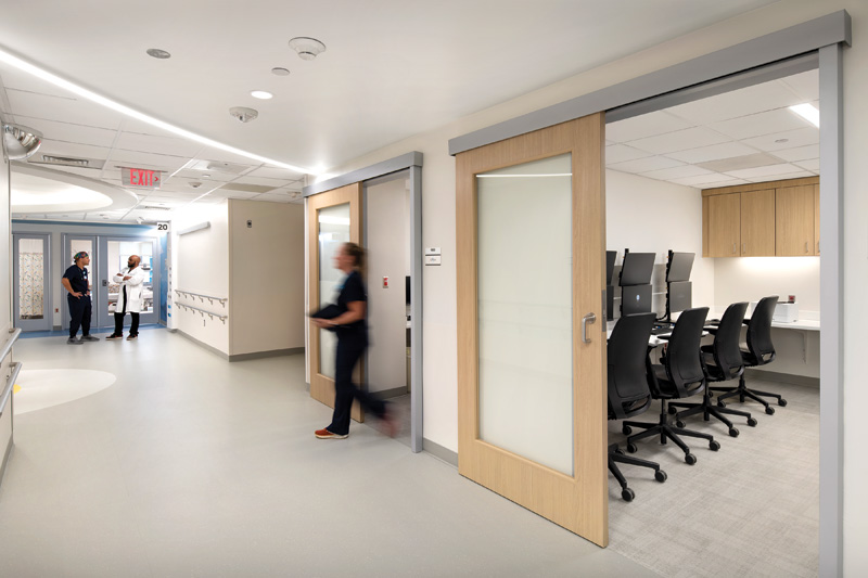 A modern hospital corridor with staff interacting, featuring a glass door office with computer workstations and accessible design elements.