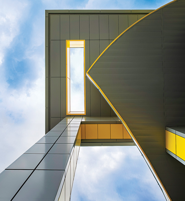 Looking up at a modern building's architectural design, featuring geometric shapes, sleek lines, and vibrant yellow accents against a cloudy sky.