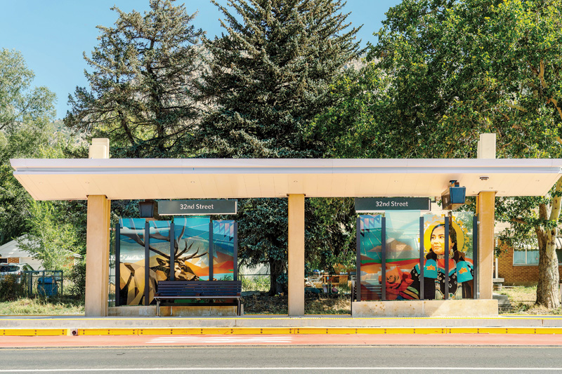 A colorful bus stop shelter with a wooden bench, surrounded by trees, featuring a sign reading "32nd Street."