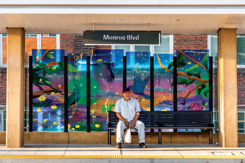 A public transit stop with a vibrant mural depicting birds and nature, featuring a bench under a sign saying "Monroe Blvd."
