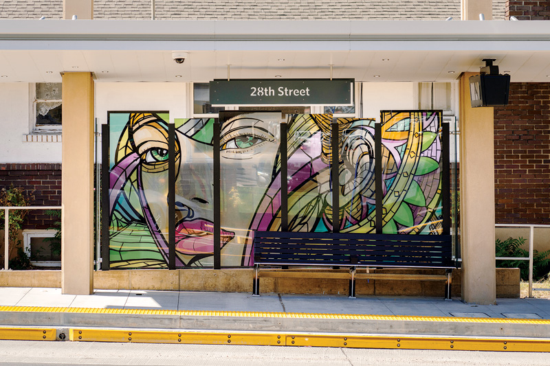 A colorful glass mural featuring faces and abstract designs at the 28th Street transit stop, with a bench for waiting passengers.