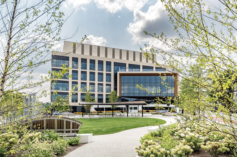 A modern office building surrounded by greenery, featuring a park-like setting with benches and a curved pathway under a cloudy sky.