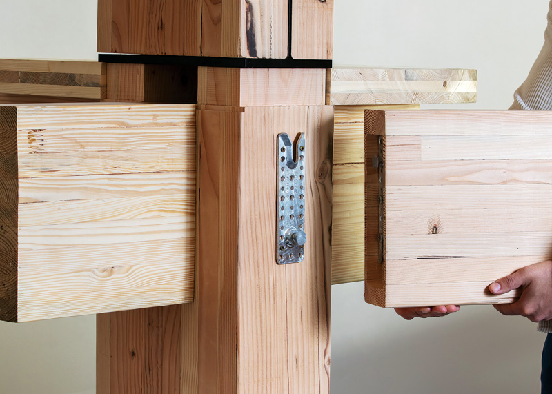A person holds a wooden beam while connecting it to a wooden structure with metal fasteners for assembly.