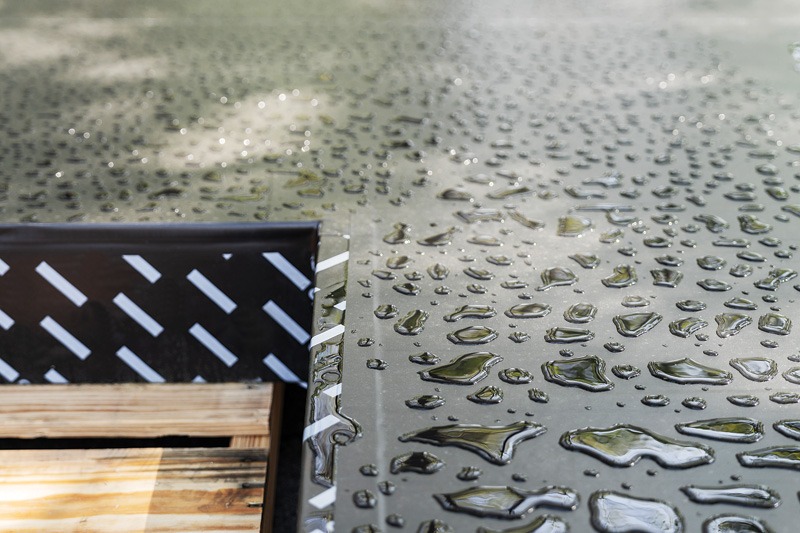Water droplets shimmer on a textured surface, with a black metal edge and wooden planks visible in the corner of the image.