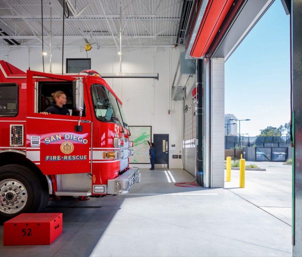 A red San Diego fire truck is positioned at the open bay of the fire station, with a map and emergency equipment visible inside.
