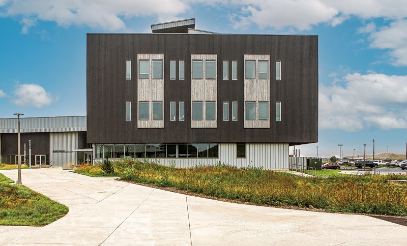 Modern building with a dark wooden facade, large windows, and a landscaped path leading to its entrance, under a partly cloudy sky.