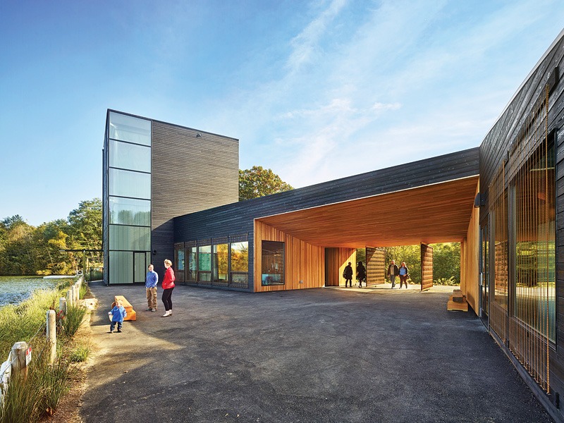 A modern wooden building with large windows, featuring families outside near a lake, under a clear blue sky.