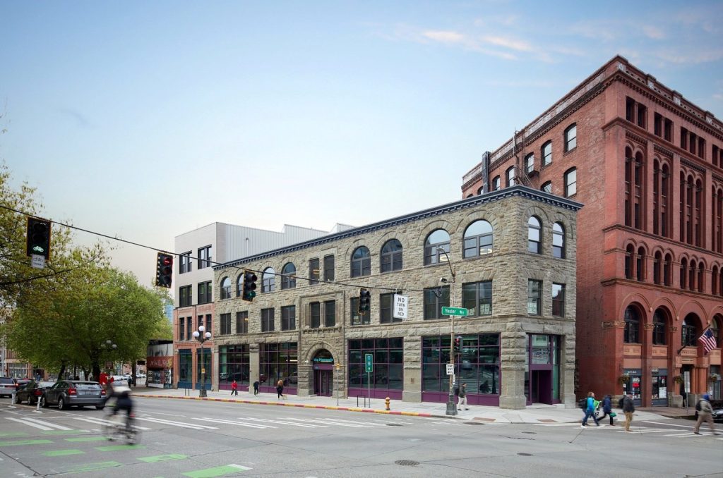 Historic stone and brick buildings at an urban intersection, featuring pedestrians, traffic signals, and greenery.