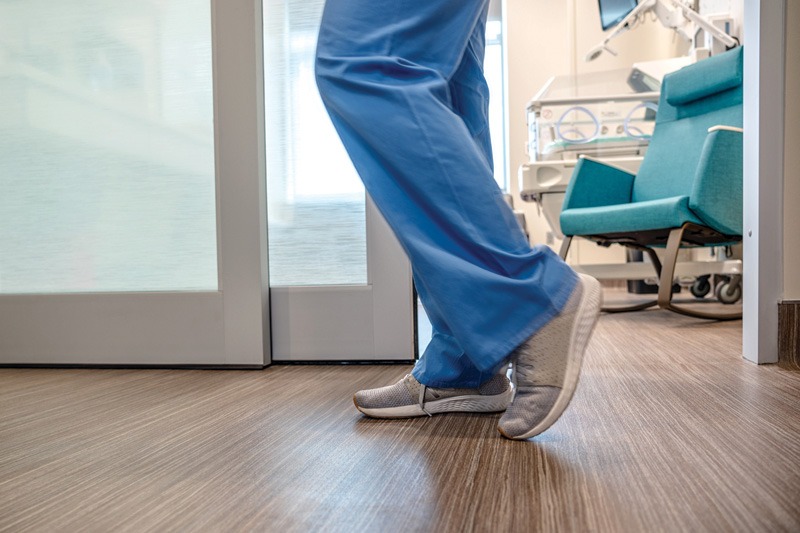 A healthcare worker in blue scrubs walks on a wooden floor, heading towards a patient room with medical equipment visible.