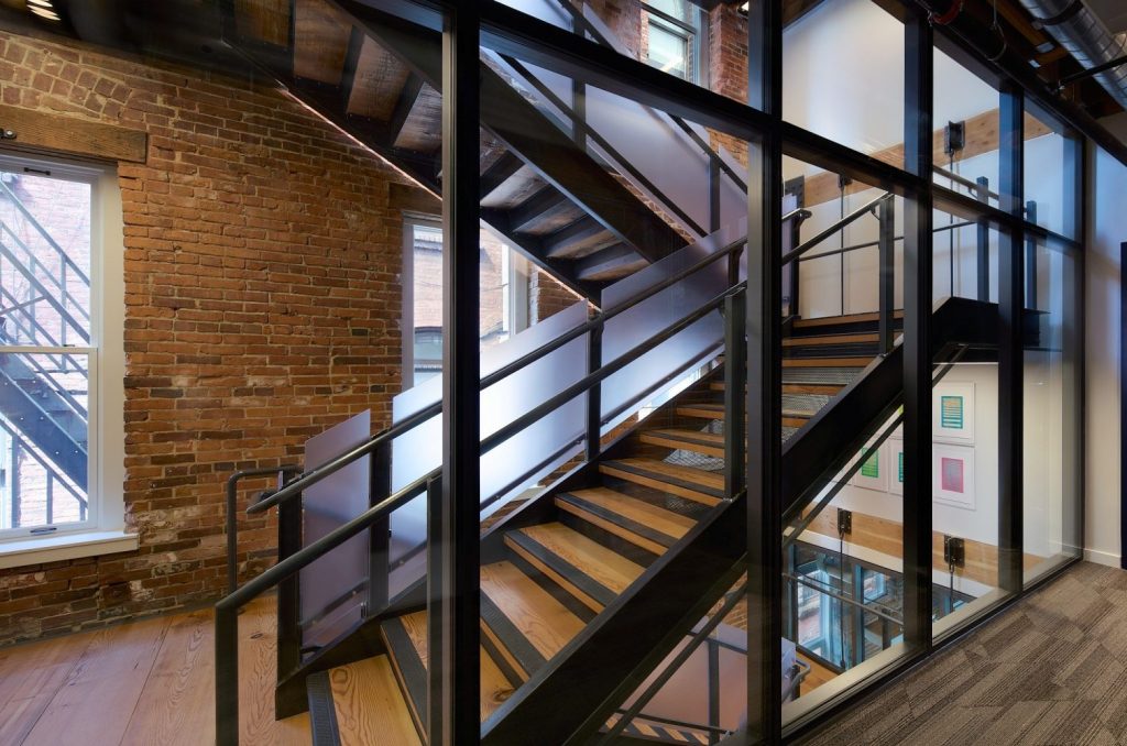 A modern staircase with wooden steps and metal railing, enclosed in glass, set against a rustic brick wall backdrop.