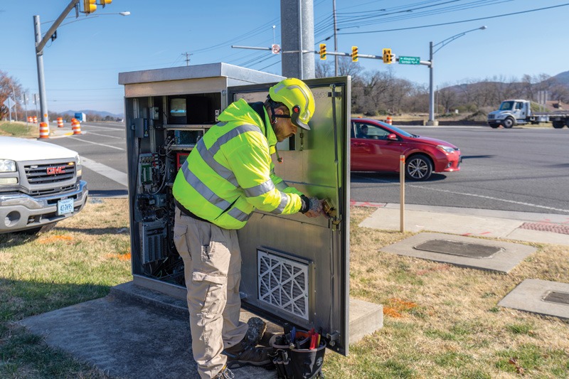 A worker in a neon yellow safety jacket repairs an electrical control box at a busy intersection with vehicles passing by.