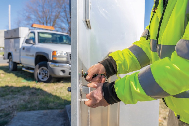 A worker in a bright yellow safety jacket uses a tool to unlock a silver cabinet, with a utility truck parked in the background.
