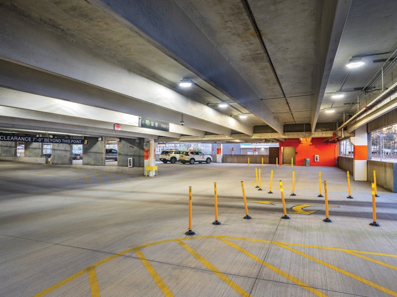 Brightly lit parking garage with yellow bollards, clear signage, and several parked cars in the background. Modern concrete architecture.