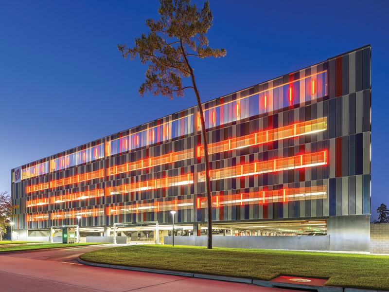 Modern parking garage facade illuminated in vibrant reds and blues, framed by a tall tree against a twilight sky.