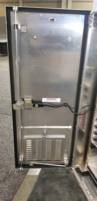 Inside view of a stainless steel vending machine door, featuring a warning label, locking mechanism, and ventilation grill at the bottom.