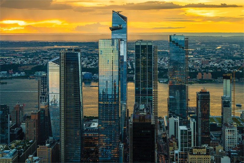 Aerial view of modern skyscrapers against a vibrant sunset over the Hudson River, showcasing reflections and urban architecture.