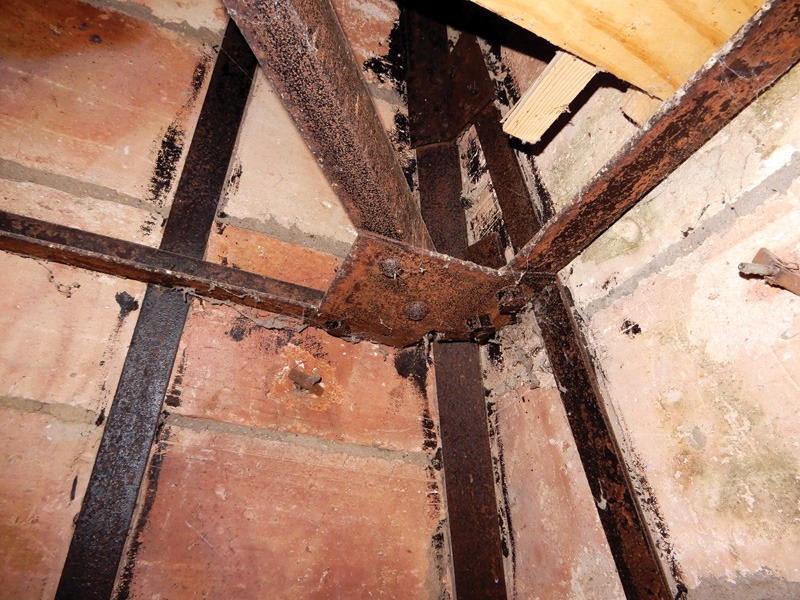 Close-up of rusted metal beams connected at a corner, with brick walls and debris visible in an unfinished space.