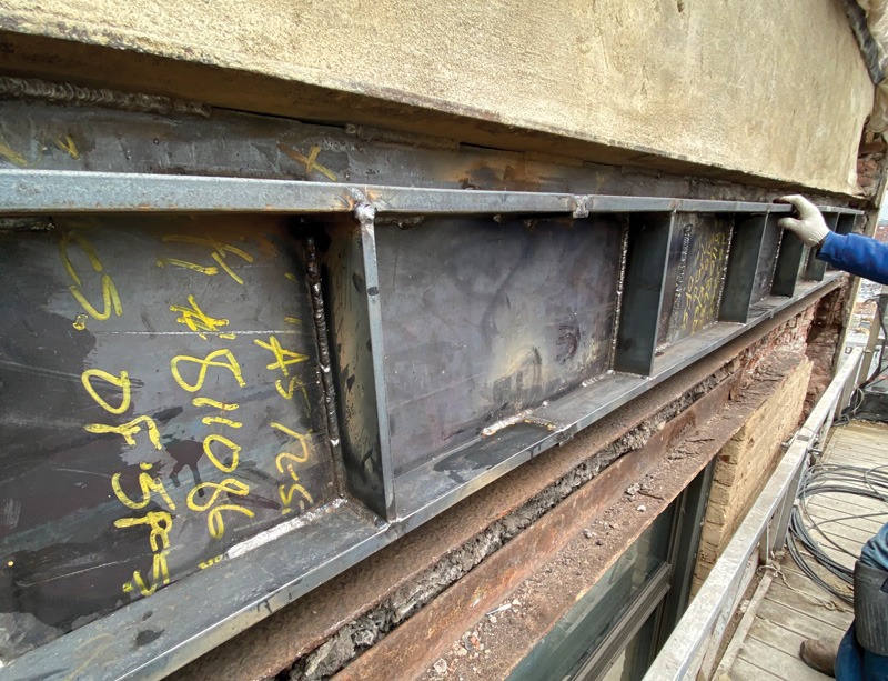 A worker inspects a steel beam with yellow markings, mounted on a building's exterior, next to a brick wall and wooden platform.