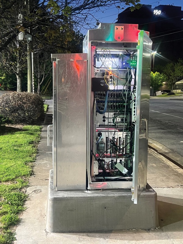 An illuminated telecommunications cabinet with open doors, revealing complex wiring and equipment, on a sidewalk at night.