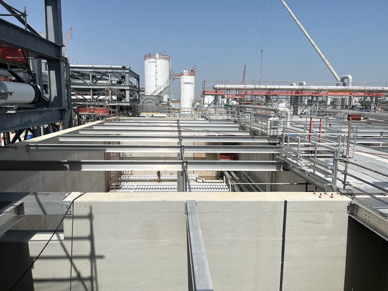 Overview of an industrial facility with metal framework, pipelines, and storage tanks under a clear blue sky.