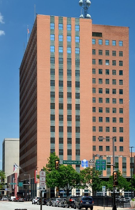 A tall, brown brick office building with a clear blue sky above, featuring traffic signs and trees in the foreground.