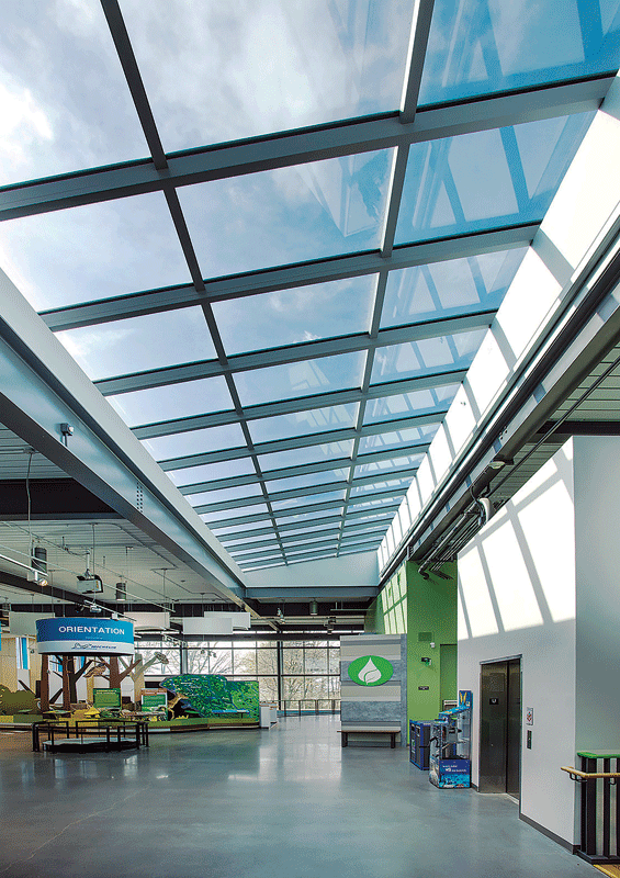 Modern atrium with a large glass ceiling allowing natural light, green and blue decor, an orientation sign, and a relaxed, welcoming atmosphere.