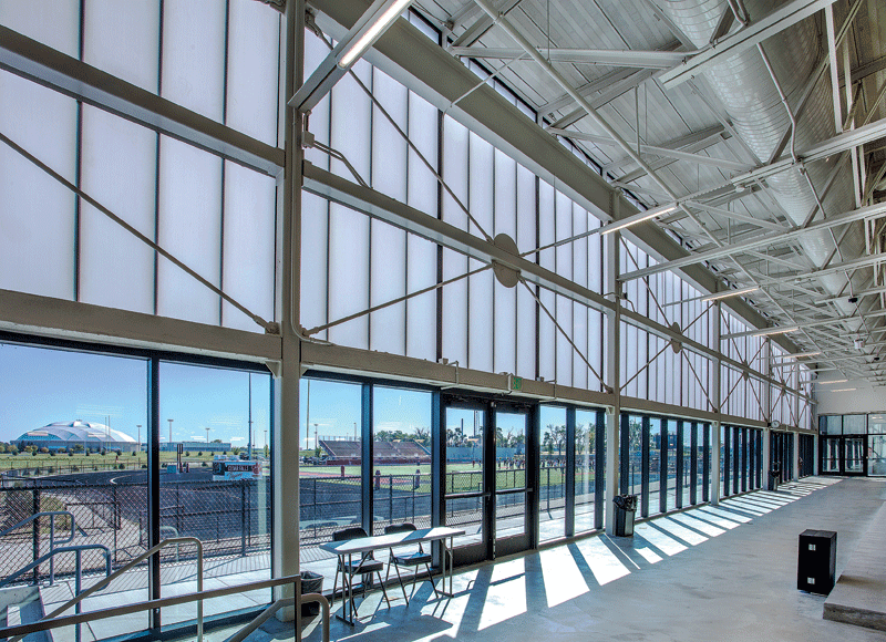 Spacious industrial interior with large windows and metal beams, overlooking a sunny outdoor sports field. Bright, open, and modern atmosphere.