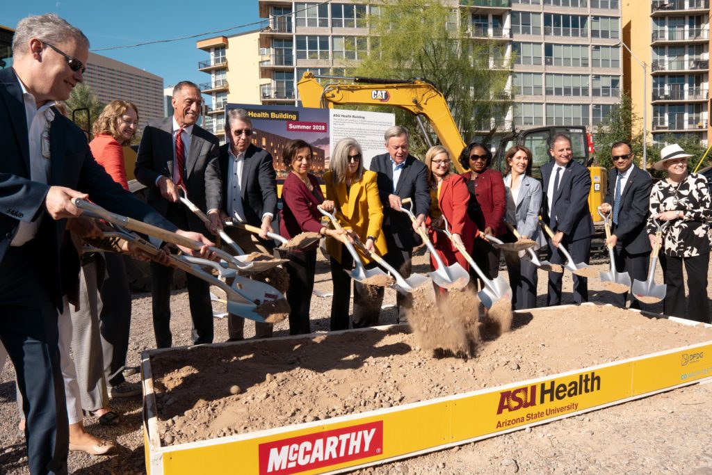 A group of people in formal attire participate in a groundbreaking ceremony, holding shovels and smiling. A construction vehicle is in the background.