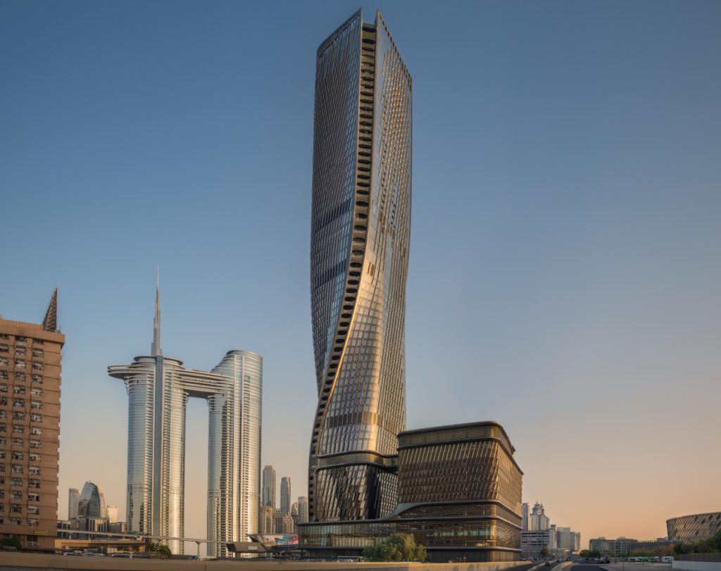 Tall, twisting modern skyscraper with reflective glass facade in a city skyline at sunset; adjacent geometric buildings create a futuristic feel.