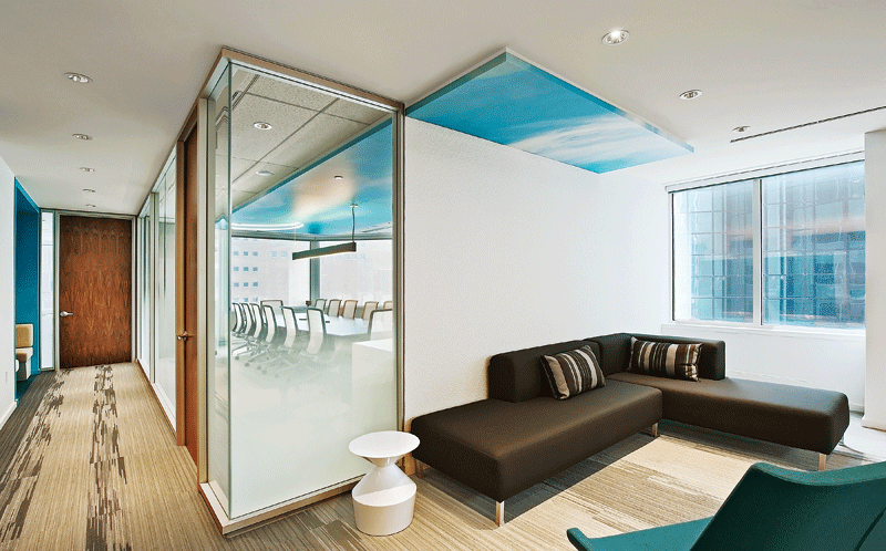 Modern office interior with a glass-walled conference room on the left, featuring white chairs around a table. A cozy seating area with a dark brown sectional sofa and a small white table is on the right. Large windows allow natural light, creating a bright and welcoming atmosphere.