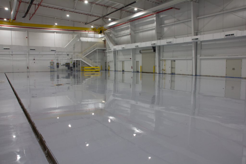 Spacious, empty industrial warehouse with shiny, polished floors, high ceilings, bright overhead lights, and white walls, conveying a pristine, clean atmosphere.