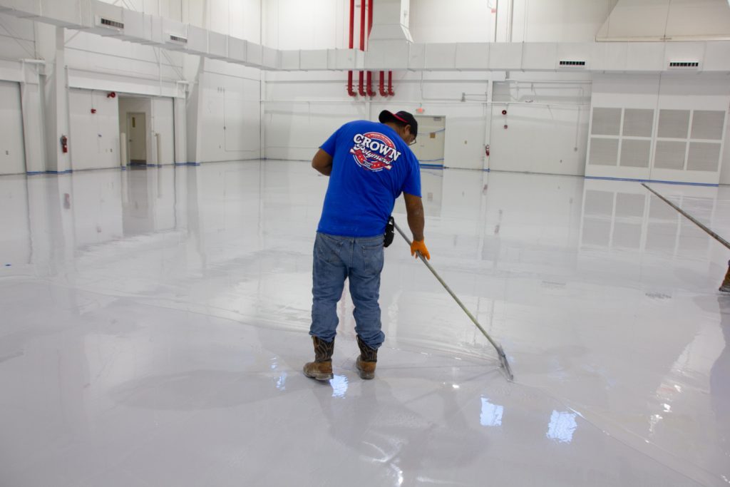 Worker in a blue shirt and jeans spreads epoxy on a large, glossy white floor in an industrial space, conveying a sense of precision and focus.