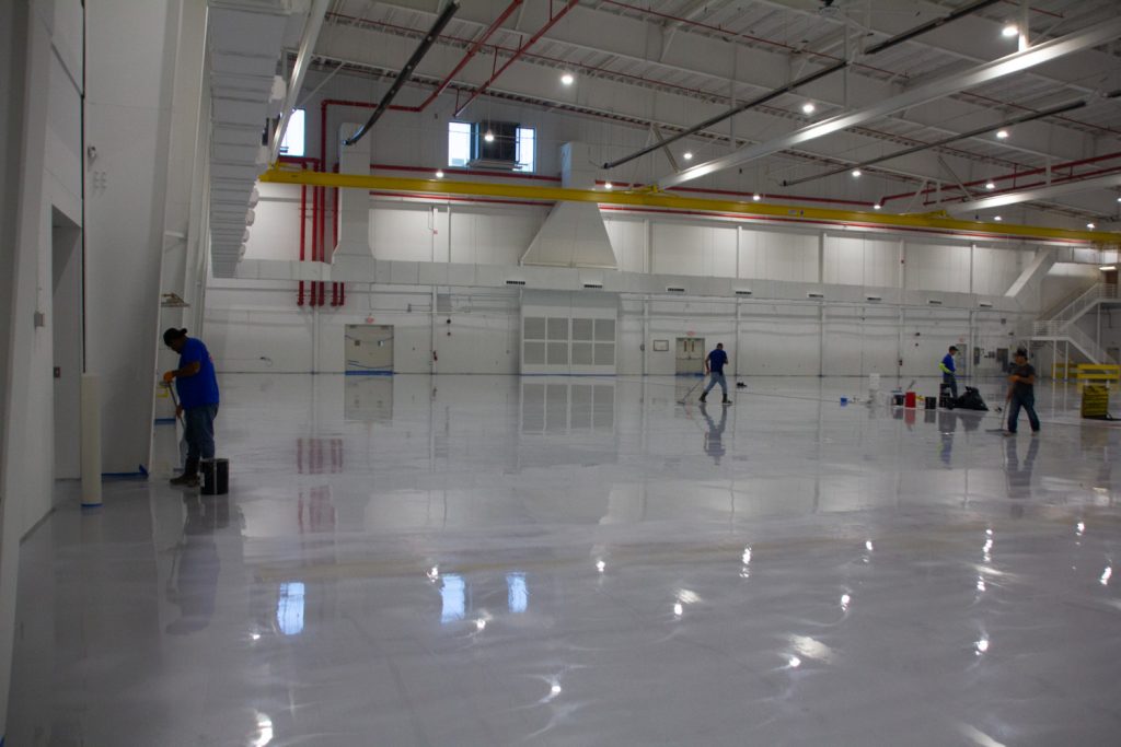 Spacious, brightly lit warehouse with a glossy floor. Several workers in blue shirts are cleaning, creating a sense of industriousness and precision.