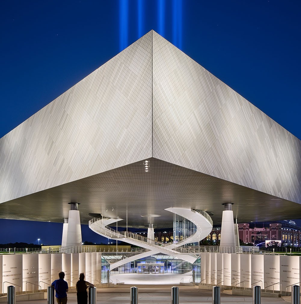 Futuristic building with illuminated facade and sharp angles under a deep blue sky. Curved walkways and two figures in the foreground convey awe.