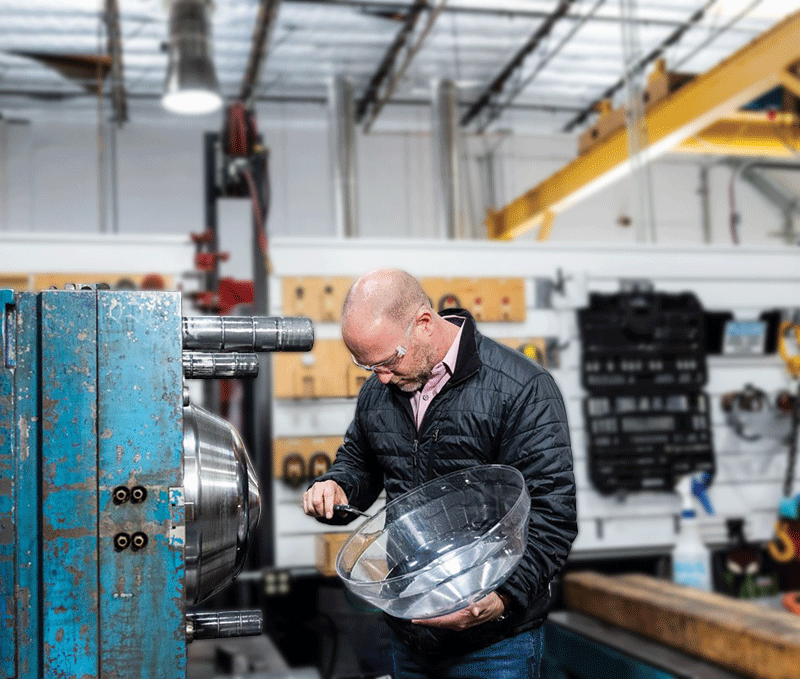 A man in a workshop inspects a clear, curved plastic part. He wears safety glasses and a black jacket. Industrial equipment and tools are visible.