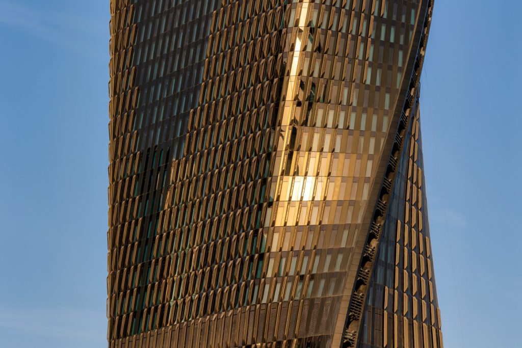 A modern skyscraper with a twisted design reflects sunlight on its golden, glass facade against a clear blue sky, conveying innovation and elegance.
