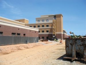A construction site with a partially completed multi-story brick and stucco building under a clear blue sky. Scaffolding and orange cones are visible.