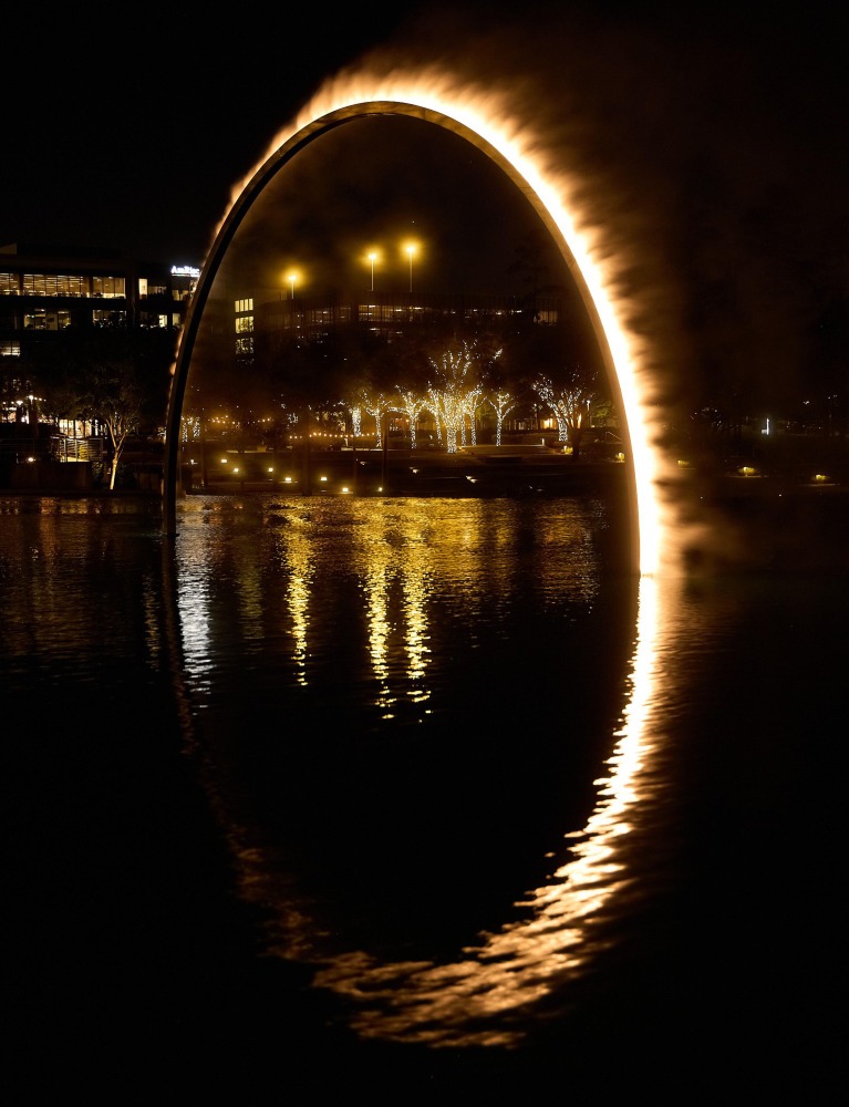 A luminous arch glows softly over a dark lake at night, reflecting golden light on the water. Twinkling trees and city lights enhance the scene's tranquility.