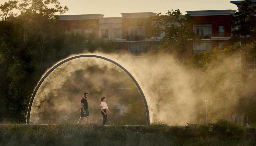 Two people walk through a circular misting arch in a park, surrounded by soft sunlight. Trees and a building are visible in the background. Calm and serene atmosphere.
