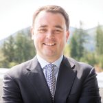 A smiling man in a suit and patterned tie stands outdoors with arms crossed. He is in front of a blurred forested mountain landscape. Bright and professional tone.