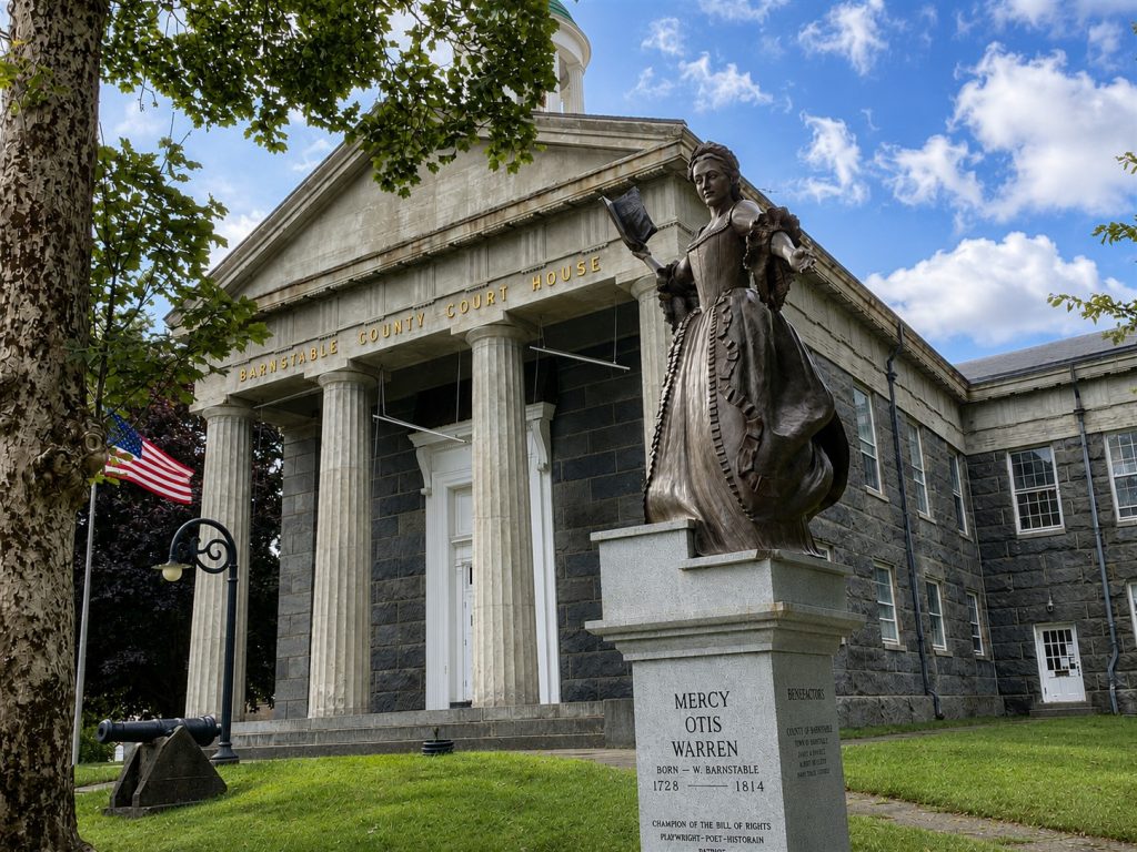 Statue of Mercy Otis Warren stands before Barnstable County Courthouse, with Greek Revival architecture, American flag, trees, and blue sky.