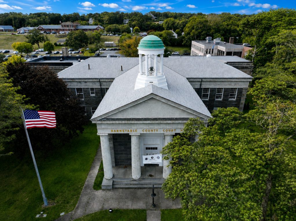 Aerial view of Barnstable County Court, featuring a neoclassical facade with columns and a green-domed cupola. A U.S. flag waves nearby, surrounded by lush trees and distant buildings under a blue sky.