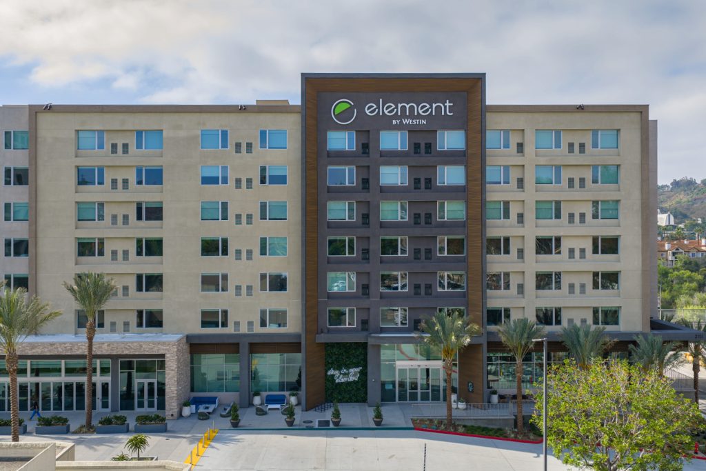 Modern six-story hotel exterior with prominent "Element by Westin" signage. Symmetrical windows, palm trees, and a sleek facade convey a welcoming and upscale atmosphere.
