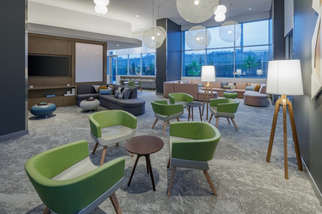 Modern lounge area with green chairs, wooden tables, and plush sofas. Large windows let in natural light, creating an inviting, airy atmosphere.