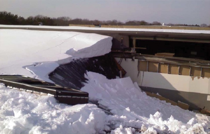 Collapsed building roof under heavy white snow, revealing damaged structures and debris. The scene conveys a sense of cold and potential danger.