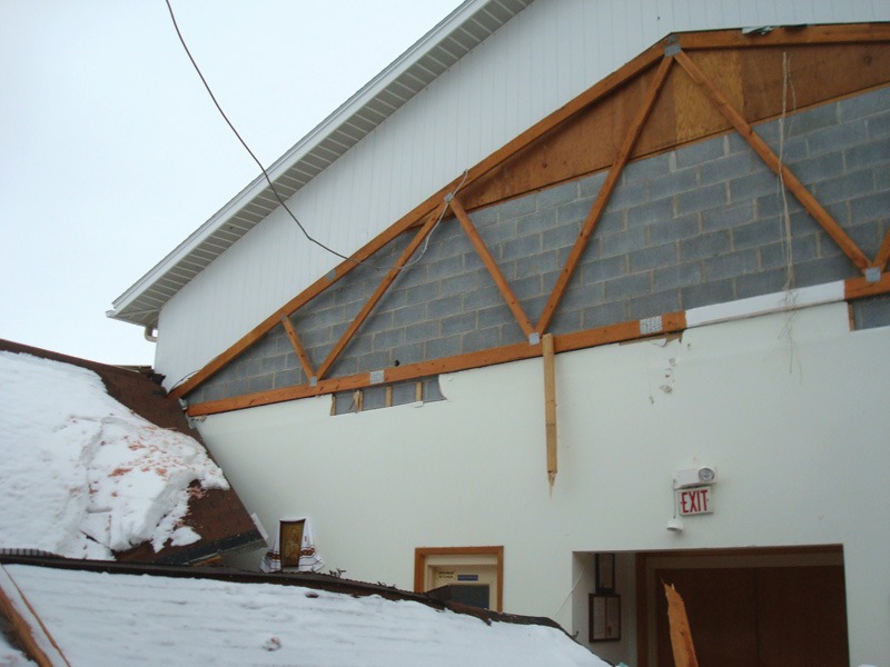Damaged building roof due to heavy snow. The ceiling and wall show structural damage, with visible wooden beams and exposed bricks. A sense of disrepair.