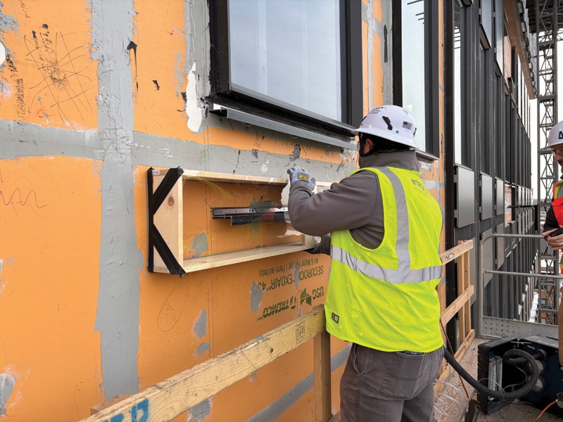 Construction worker in a hard hat and neon vest measures a window frame on an orange building facade. Another worker observes. Overcast weather.
