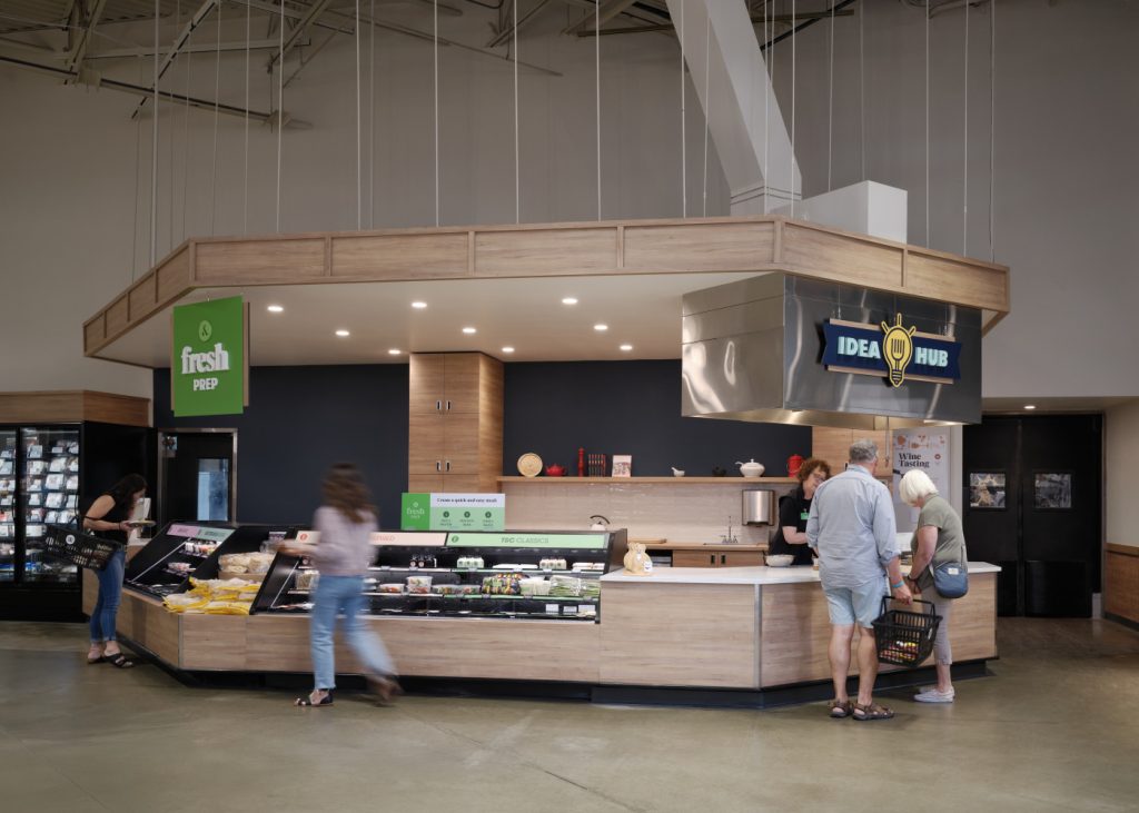Market interior with people shopping at a deli counter. The area is labeled "fresh prep" and "idea hub," with a display of food items in a modern setting.