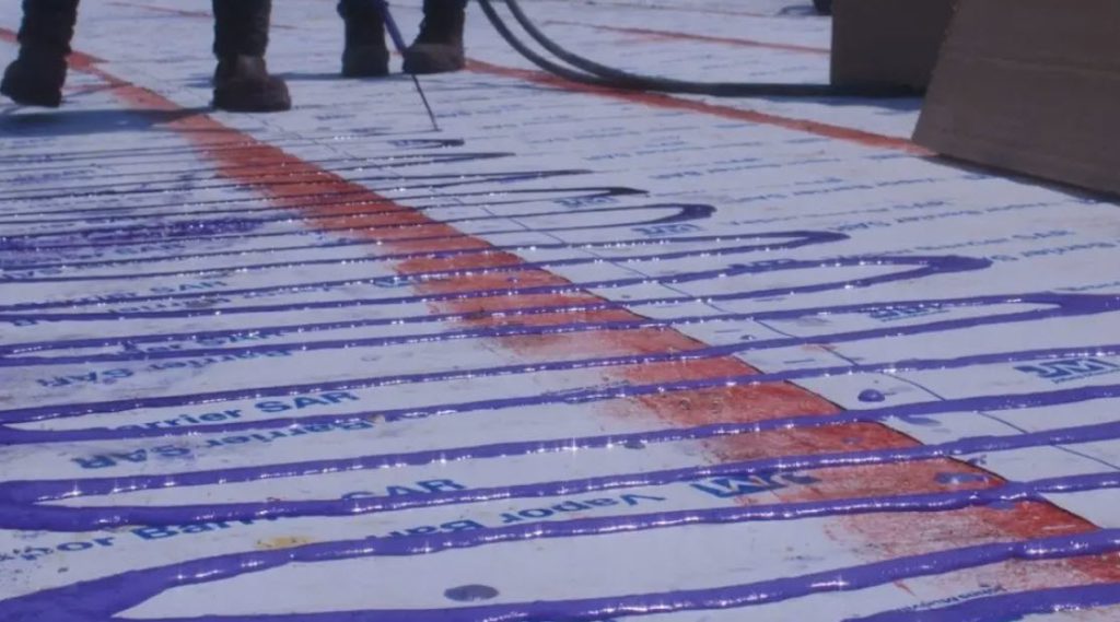 Workers apply purple waterproofing liquid on a construction surface marked with orange grid lines and text. The scene conveys a sense of precision.