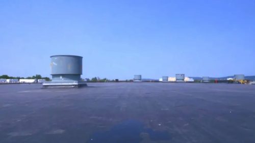 A flat, expansive rooftop under a clear blue sky features several large ventilation units. The scene is calm with distant trees at the horizon.